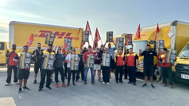 A group of people holding signs and flags stands in front of DHL-branded trucks, in a union rally