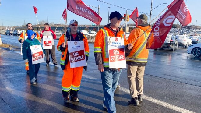 A group of workers from Unifor Local 100, identifiable by their signs and flags, are participating in a strike. They are wearing high-visibility safety gear and are marching in a parking area under clear skies, conveying a sense of solidarity and purpose.