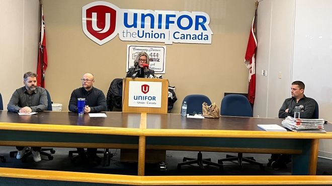 A woman speaks at the podium while three men sit in chairs. There is a large Unifor logo banner behind her and two red Unifor flags on each side of the room.