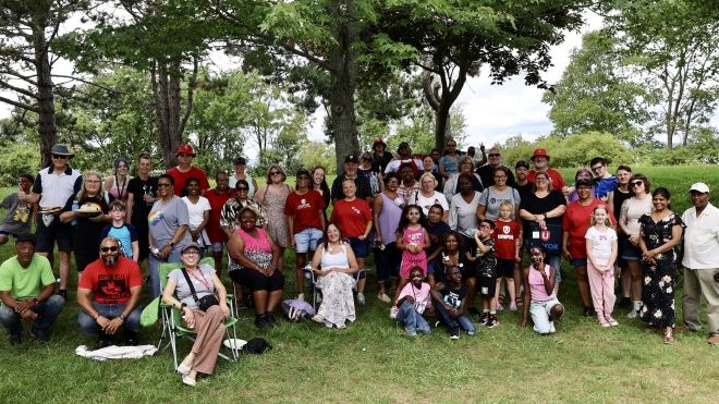 A large group pose outside on the grass in front of green trees