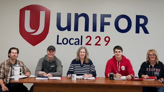 Five members of the Unifor Local 229 paramedic bargaining committee are seated at a table in front of a wall with the Unifor logo and “Local 229” text. They are facing the camera, with bargaining materials, coffee mugs, and folders in front of them.