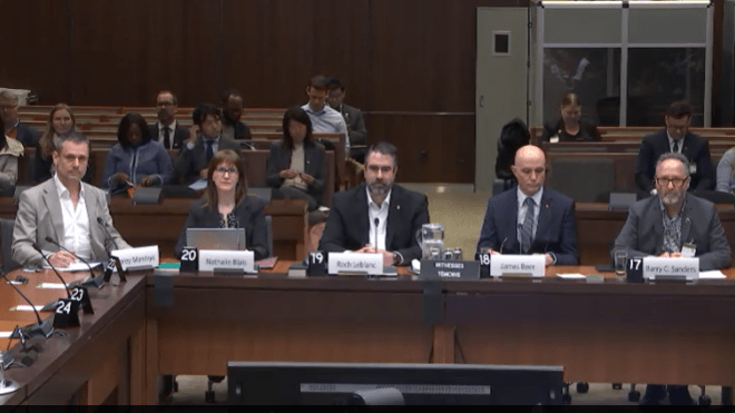 Five people sitting in a meeting square table and others listening in the background 
