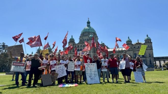 Gavin McGarrigle speaking from a glass podium with Shaun Bhoondpaul standing behind him. The BC legislature building with a giant Canadian flag is in the background. 