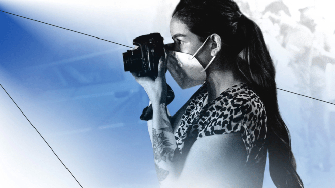 A woman wearing a mask looking through the lens of a camera.