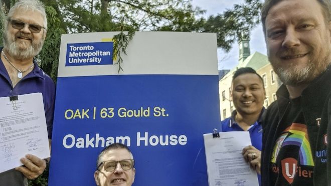 Four people posing beside an Oakham House sign holding documents."