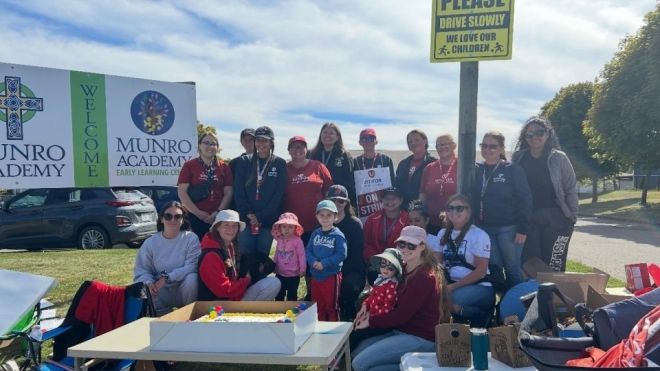  A group of women and three children, some wearing red and holding "On Strike" signs, gather outside near Munro Academy banners and a "Please Drive Slowly, we love our children" sign on a sunny day, at a picket line. 