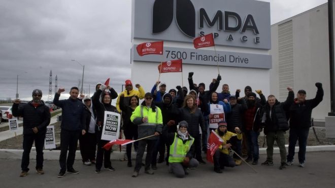 A group of individuals waving flags and raising fists in a demonstration outside the MDA Space building at 7500 Financial Drive, with some holding signs that read "MDA ON STRIKE FOR DECENT PENSIONS NOW NO CONTRACT NO WORK" and "Unifor Union on strike."