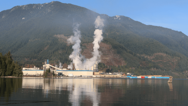 A shoreline pulp mill with smoke rising out of smokestacks and a treed mountain in the background.