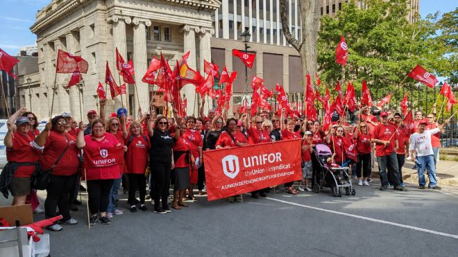 Unifor members gather outside a columned building, waving red union flags and holding a banner reading ‘A union defending rights for everyone.