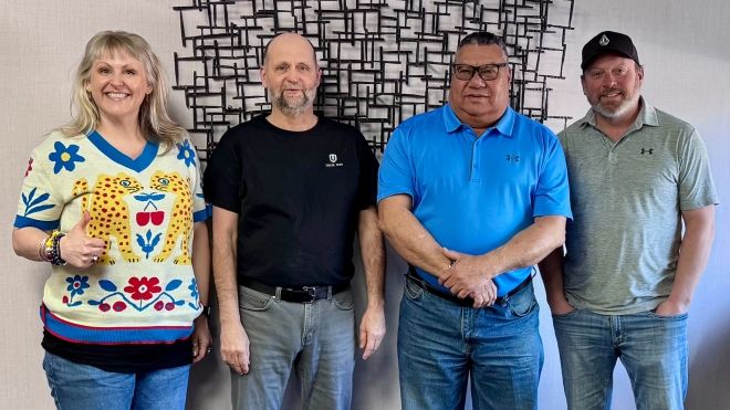 Four people standing near a wall indoors. One gives the "thumbs up" sign.