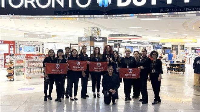 Thirteen women dressed in all black hold up red Unifor placards in front of a Toronto Duty Free store