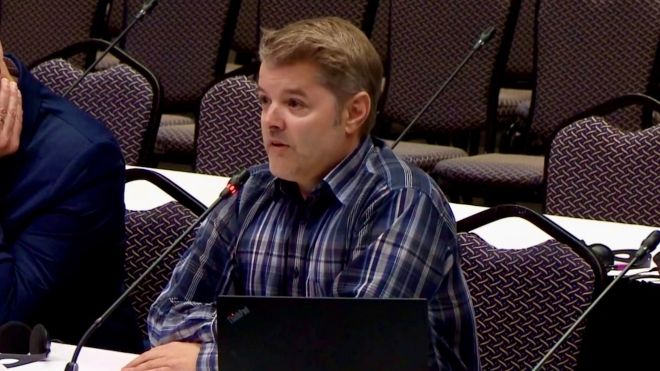 Two men sit at a table with a microphone while one speaks.