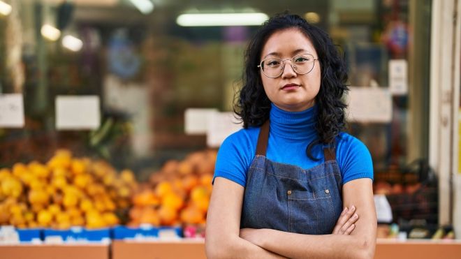 Young Chinese woman worker standing with arms crossed at local grocery store produce section.