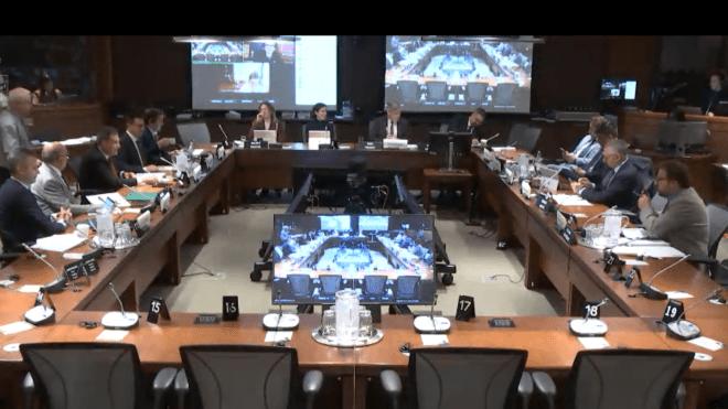 Hollow square table configuration with people seated and large television screens in the background.