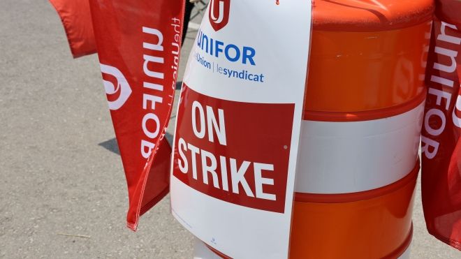 A large orange construction cone draped with Unifor flags and an sign reading "On Strike"