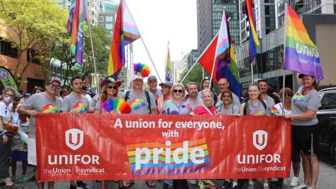 Unifor members, leadership and staff smile at Toronto Pride in front of a Unifor banner.