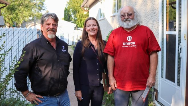 “Tracy Schmidt standing between two men on a doorstep. The men are wearing shirts with Unifor logos.”