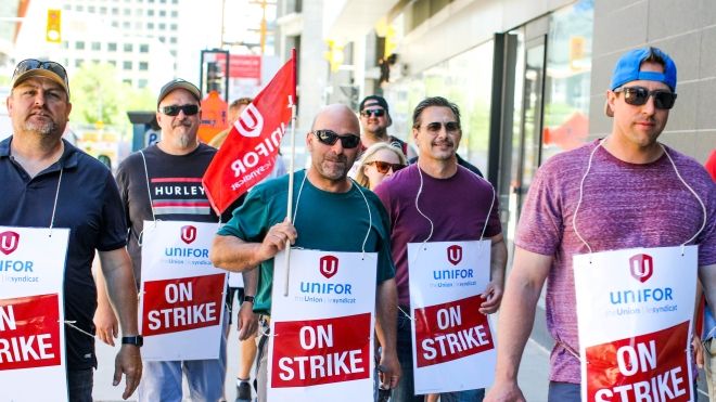 Row of six striking Manitoba Hydro workers walking on the sidewalk wearing On Strike signs