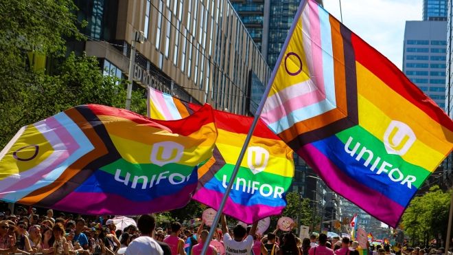 A large crowd of people at a parade with three large Unifor Pride Flags in the foreground.