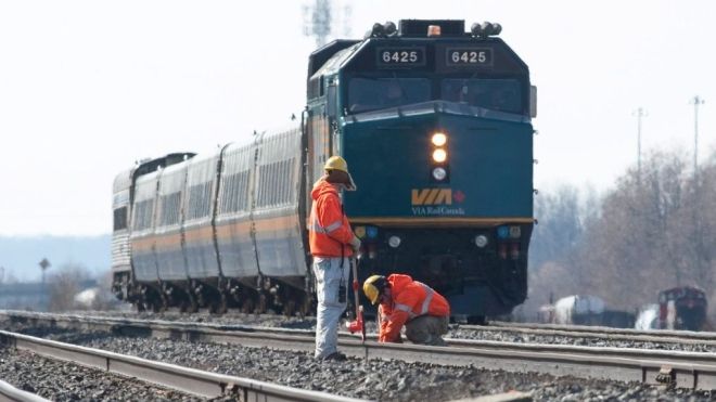 Two workers in safety gear inspect railway tracks as a VIA Rail train approaches in the background on a sunny day.