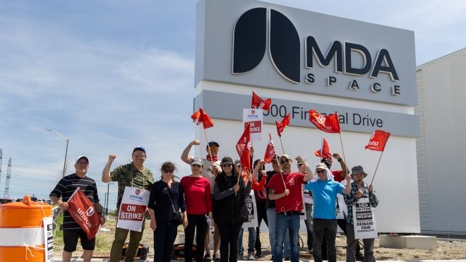 A group of people holding up Unifor flags and place cards outside in front of a MDA Space exterior sign