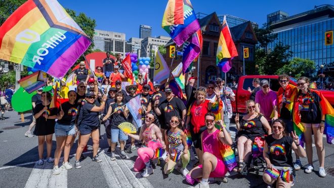 A large group at the Toronto Pride parade waving Pride flags in front of a colourful Unifor float
