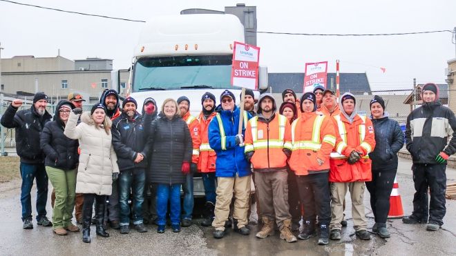 Lana Payne among a line of approximately 20 workers standing on a picket line holding fists in the air in front of a white truck