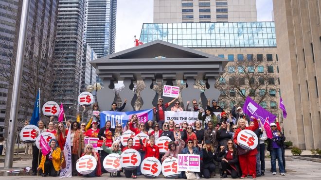 A large group of people pose outside a building, holding signs and banners advocating for closing the gender pay gap and wage equality.