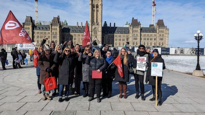 A group of people stand in front of Parliament Hill holding signs calling for pharmacare
