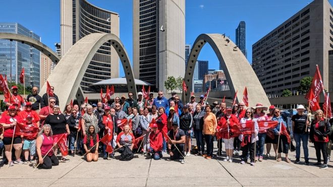 A huge group shot in front of Nathan Phillips Square Toronto. Most are wearing red and waving red Unifor flags