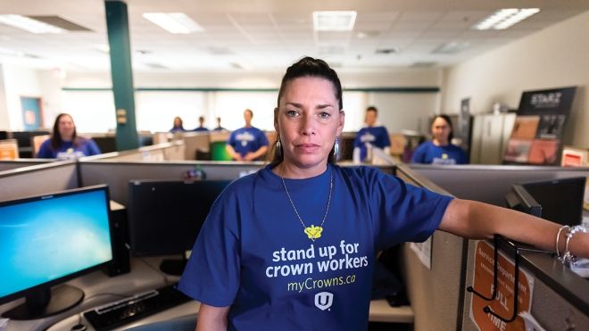 Woman standing in cubicle in foreground with Stand Up For Your Crowns t-shirt and other office workers in the background.