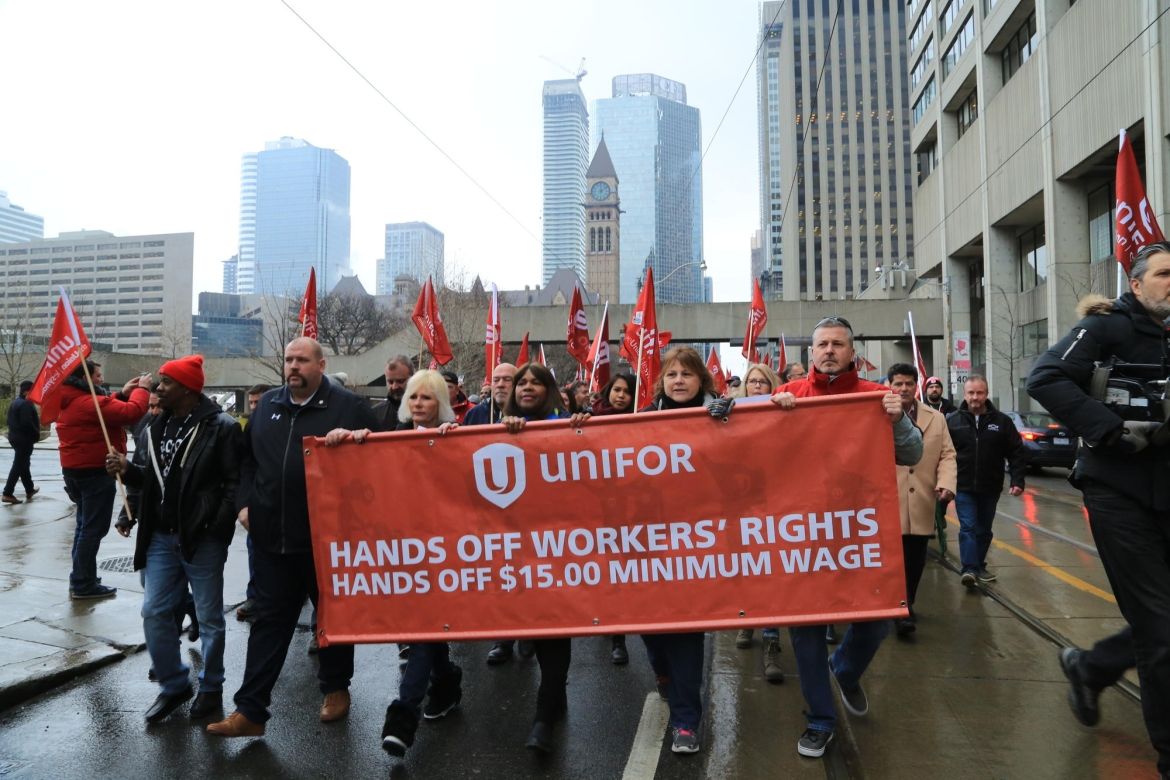 Unifor members march behind a banner reading &quot;Hand off workers' rights. Hands off $15 minimum wage.&quot;