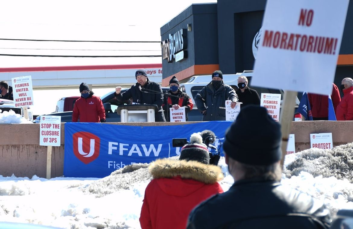 Keith Sullivan speaking from a podium outdoors with person holding 'No Moratorium' sign in foreground