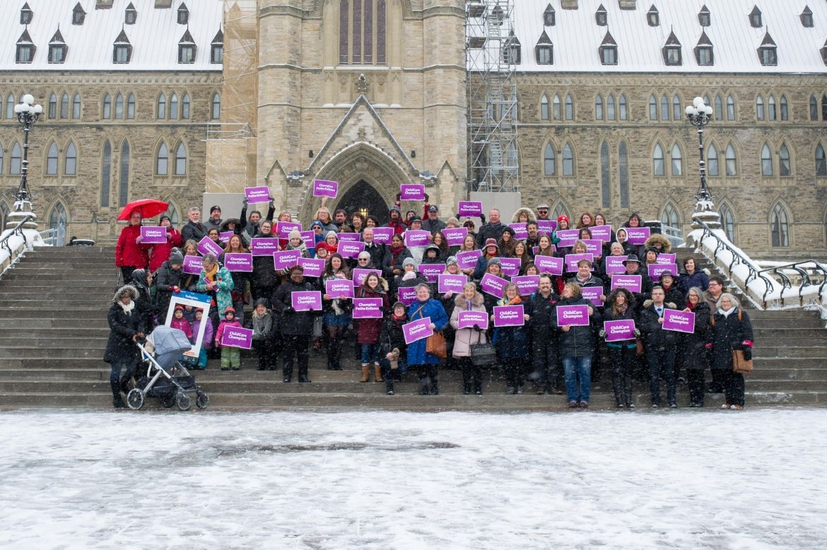A large group of activists on the steps of Parliament Hill holding purple Child Care Champion signs.