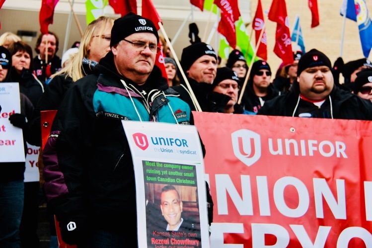 Members of Local 597 in matching toques at a rally, in the foreground one member wears a sign denouncing their employer's CEO.