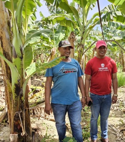 Man in red shirt and man in blue shirt with banana trees in background