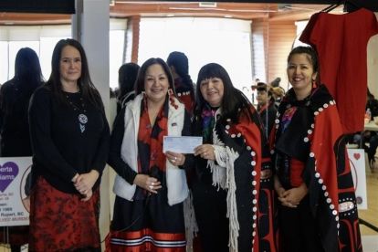 Four women, two wearing ribbon skirts and two wearing blankets with Indigenous styling pose holding a cheque.