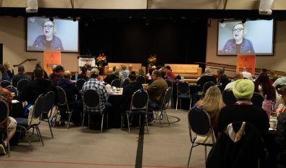 Two screens seen from the back of a room, a women's face displays on screen
