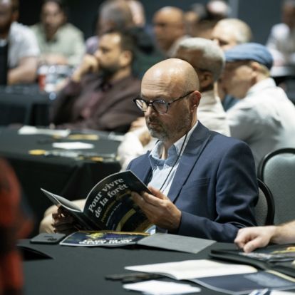 Seated man reading a document with other seated at round tables behind him.