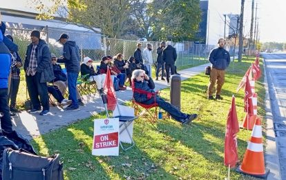 A group of picketers sitting roadside near orange pillions and "on strike" signs