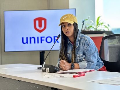 A women wearing a yellow hat sits at a table in front of a screen with a Unifor logo
