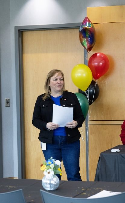 A women holding a paper standing in front of a table with a flower vase, ballons in behind