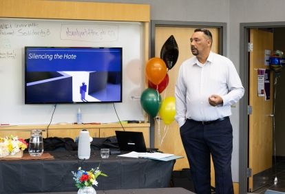 A man speaking, a monitor and balloons in the background