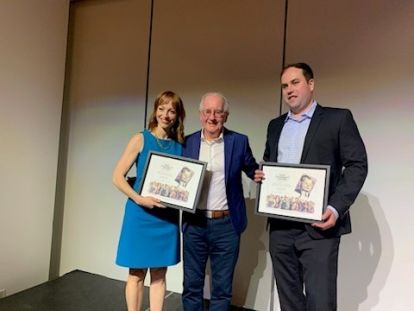 A woman wearing a blue dress and a man wearing a dark suit, both holding awards, stand next to a man in a blue suit.