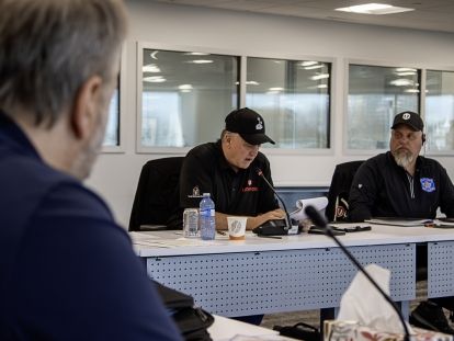 A seated man speaks into a microphone. Another man is seated beside him and in the foreground.