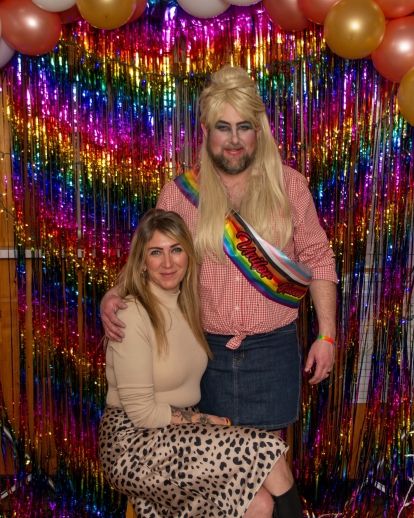 A woman sits on a stool, a drag queen stands beside her.