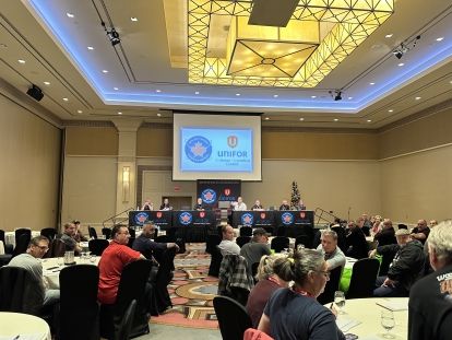 people sitting at tables looking front at a screen with a Unifor Skilled Trades Logo a red flag with a blue circle around it.