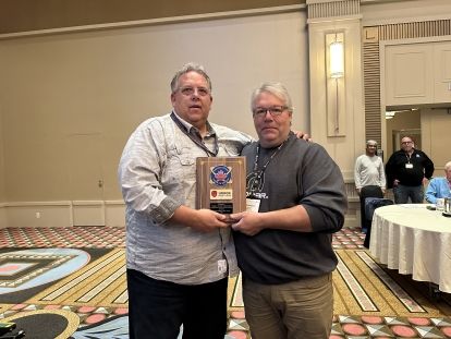 Two man standing arms around each other holding a plaque