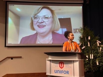 A women speaking on a large screen on Zoom and a women stands in front at a podium.
