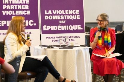 Two women speaking into mics sit table side in front of IPV banners